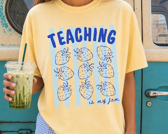 May include: A pale yellow short-sleeve t-shirt featuring the blue text TEACHING is my jam and a graphic pattern of nine blue strawberry illustrations arranged in a grid over vertical light blue stripes on the front of the garment.
