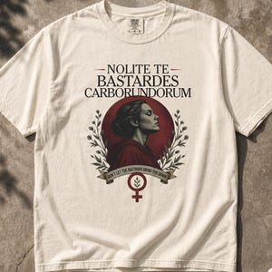 Könnte beinhalten: Ein elfenbeinfarbenes Kurzarm-T-Shirt mit einem Grafikdruck einer Person im Profil in einem roten Kreis, flankiert von botanischen Zweigen. Der Text lautet NOLITE TE BASTARDES CARBORUNDORUM und DON'T LET THE BASTARDS GRIND YOU DOWN, mit einem Frauensymbol.