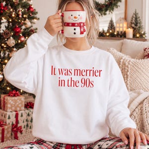 Peut inclure: Un sweat-shirt blanc à manches longues avec le texte rouge It was merrier in the 90s imprimé sur le devant. Le vêtement est associé à un bas de pyjama à carreaux et à une tasse en céramique en forme de bonhomme de neige avec une écharpe rouge.