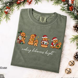 May include: A moss green crewneck t-shirt featuring a graphic of four gingerbread characters engaged in various activities, with the text "making behaviors bright" written in white script below the illustration.