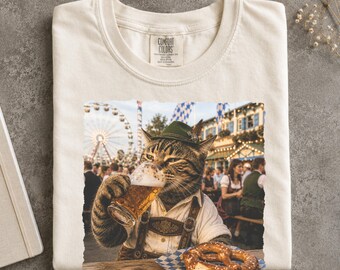 May include: An ivory t-shirt featuring a graphic print of a tabby cat wearing a green alpine hat and lederhosen, holding a glass of beer at a fair. The cat sits at a table with a pretzel. The shirt has a Comfort Colors brand label at the neck.