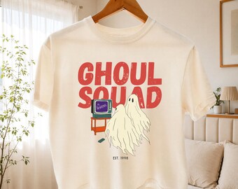 May include: An ivory short-sleeve t-shirt featuring a graphic print with the red text GHOUL SQUAD above two white ghosts watching a small television on a stand. The television screen displays the text THE PARANORMAL and the text EST. 1998 is below.