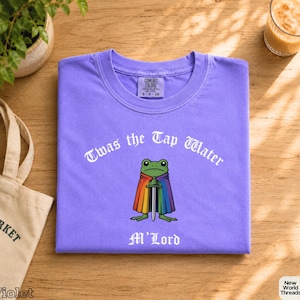 Pode incluir: Uma t-shirt violeta com um gráfico de um sapo verde a usar uma capa com as cores do arco-íris e a segurar uma espada. O texto "Twas the Tap Water" está impresso a branco acima e "M'Lord" está impresso a branco abaixo.