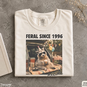 Pode incluir: Uma t-shirt cor de marfim com o texto FERAL SINCE 1996 sobre um gráfico de um guaxinim com óculos de sol e um chapéu de festa, sentado na parte de trás de um veículo a segurar um cupcake. O nome nicole está impresso em letra cursiva.