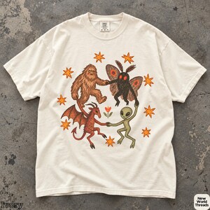 Può includere: Una t-shirt a maniche corte color avorio con una grafica centrale che raffigura un Bigfoot marrone, una creatura falena nera, un demone alato rosso e un alieno verde che si tengono per mano in cerchio, circondati da stelle arancioni e un fiore.