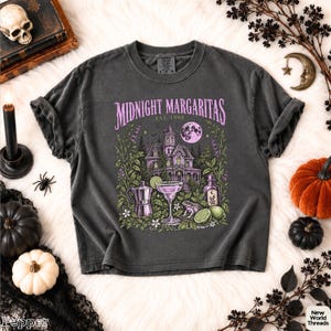 May include: A charcoal gray t-shirt featuring a graphic print with the text Midnight Margaritas Est. 1998. The design includes a Victorian-style house, a full moon, a cocktail glass, a coffee pot, a bottle, a frog, and lime slices in purple and green.