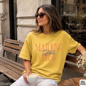 May include: A mustard yellow short-sleeve t-shirt featuring the text "MAHJONG Girls CLUB" printed on the front. The word "MAHJONG" and "CLUB" are in a bold red serif font, while the word "Girls" is in a blue cursive script font.