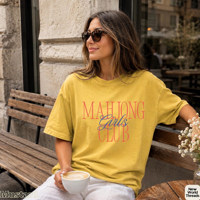 May include: A mustard yellow short-sleeve t-shirt featuring the text "MAHJONG Girls CLUB" printed on the front. The word "MAHJONG" and "CLUB" are in a bold red serif font, while the word "Girls" is in a blue cursive script font.