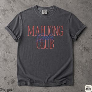 May include: A charcoal grey short-sleeve t-shirt with a crew neckline. The front features the text MAHJONG in large coral letters, the word Girls in blue cursive script, and the word CLUB in large coral letters centered on the chest.