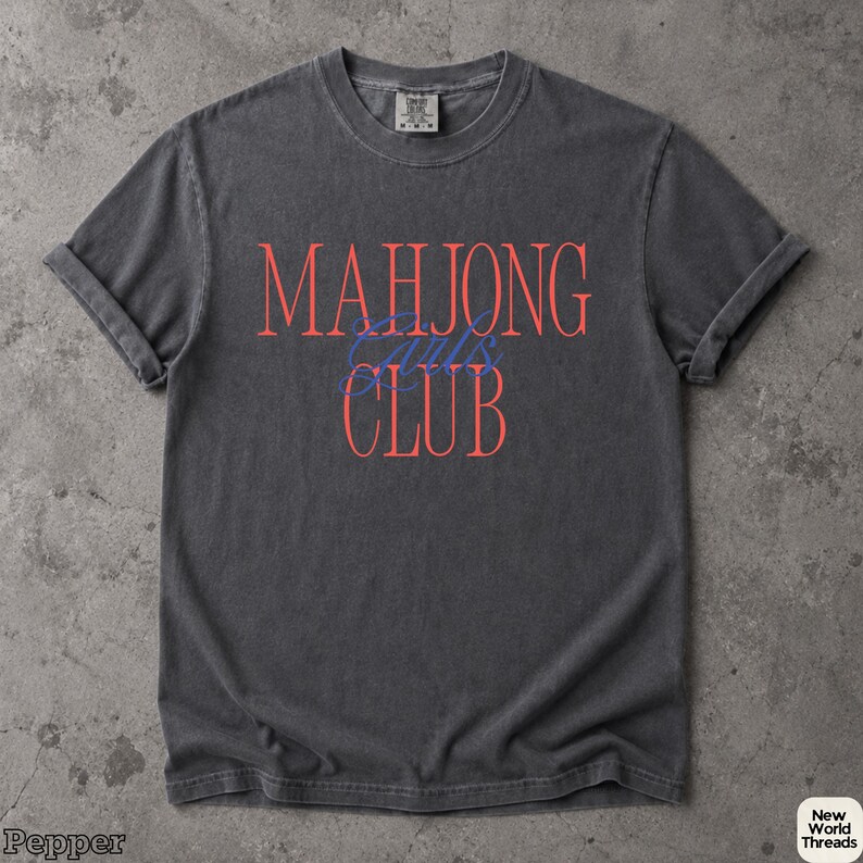 May include: A charcoal grey short-sleeve t-shirt with a crew neckline. The front features the text MAHJONG in large coral letters, the word Girls in blue cursive script, and the word CLUB in large coral letters centered on the chest.