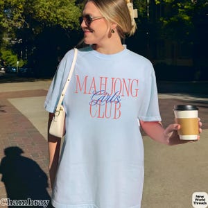 May include: A light blue short-sleeve t-shirt featuring the text "MAHJONG" in large orange letters, "Girls" in blue cursive script, and "CLUB" in large orange letters. The shirt has a crew neckline and a relaxed fit, worn with a white shoulder bag.