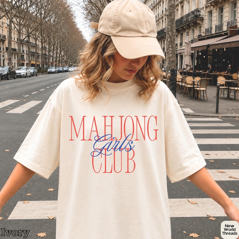 May include: An ivory short-sleeve t-shirt featuring the text "MAHJONG" in red, "Girls" in blue script, and "CLUB" in red. The shirt is paired with a solid beige baseball cap. The text "Ivory" and "New World Threads" appear in the corners.
