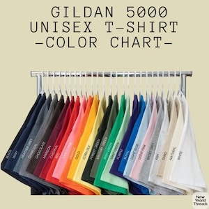 May include: A Gildan 5000 unisex t-shirt color chart displaying a wide variety of short-sleeved shirts hanging on a metal rack. The shirts are arranged in a spectrum of colors including black, navy, red, orange, yellow, green, blue, pink, gray, and white, with each color labeled clearly below the fabric.