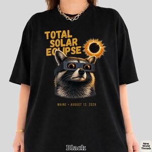 May include: A black t-shirt featuring a graphic of a raccoon wearing eclipse glasses, looking at a solar eclipse. The text reads TOTAL SOLAR ECLIPSE and MAINE AUGUST 12, 2026. The design uses yellow and orange tones against the black fabric background.