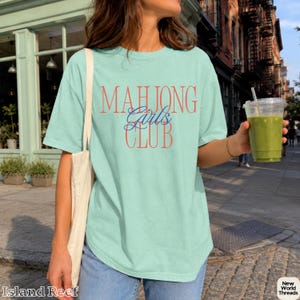 May include: A light mint green short-sleeve t-shirt featuring the text "MAHJONG" in large red letters, "Girls" in blue cursive script, and "CLUB" in large red letters. The shirt is paired with a plain white tote bag and blue denim jeans.
