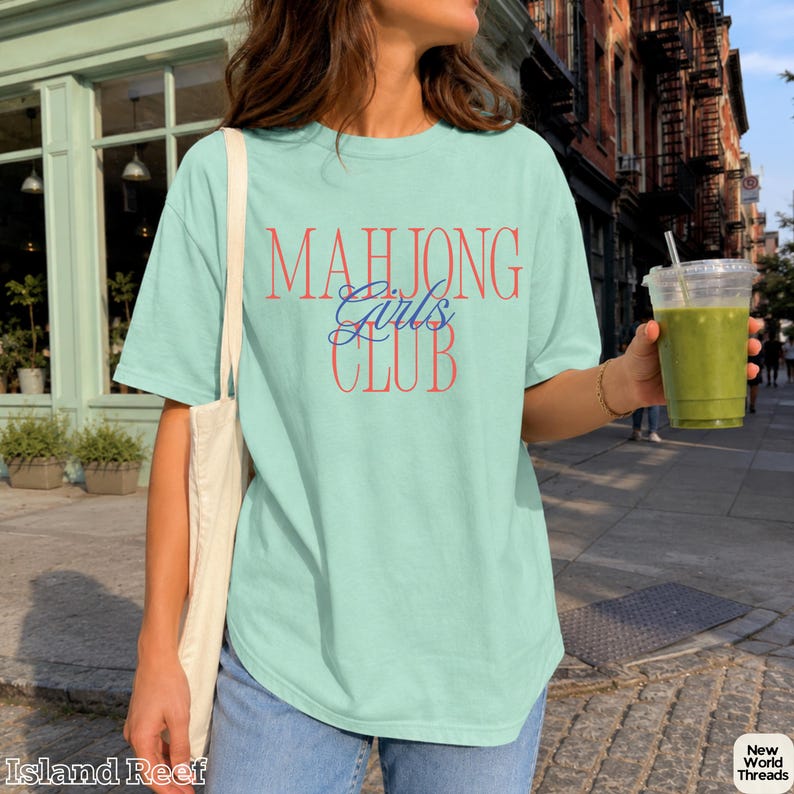 May include: A light mint green short-sleeve t-shirt featuring the text "MAHJONG" in large red letters, "Girls" in blue cursive script, and "CLUB" in large red letters. The shirt is paired with a plain white tote bag and blue denim jeans.