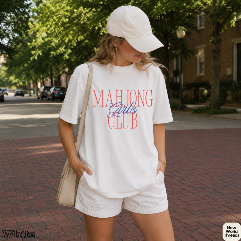 May include: A white short-sleeved t-shirt featuring the red text "MAHJONG" and "CLUB" with the blue script text "Girls" in the center. The shirt is paired with white shorts, a beige shoulder bag, and a plain white baseball cap.