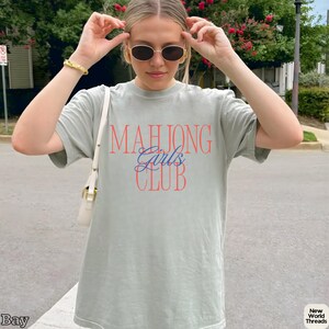 May include: A sage green short-sleeve crewneck t-shirt featuring the text "MAHJONG Girls CLUB" printed on the front. The word "MAHJONG" and "CLUB" are in a bold red serif font, while the word "Girls" is in a blue cursive script font.
