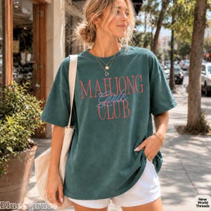 May include: A dark teal short-sleeve t-shirt featuring the text "MAHJONG" in red, "Girls" in blue script, and "CLUB" in red. The shirt is paired with white shorts and a beige canvas tote bag, accessorized with a layered gold necklace.