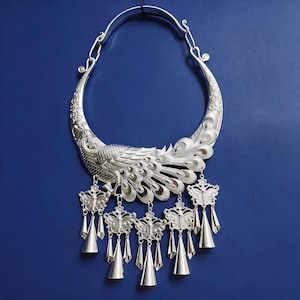 Può includere: Una collana rigida in metallo color argento con un intricato design a pavone e dettagli di piume incise. Il pezzo include cinque ciondoli a forma di farfalla, ognuno rifinito con un piccolo pendente liscio a forma di cono alla base.