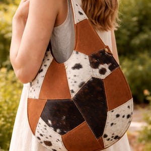 Może przedstawiać: Patchworkowa torba na ramię o unikalnym designie. Torba posiada brązowe, białe i czarne panele ze skóry bydlęcej. Torba ma zaokrąglony kształt i pojedynczy pasek na ramię. Torba jest trzymana przez osobę ubraną w białą sukienkę.