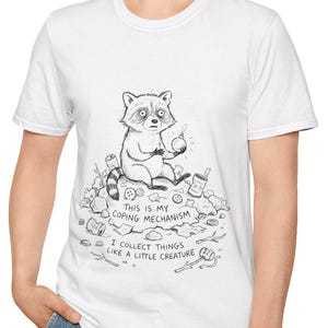 May include: White t-shirt featuring a black and white illustration of a raccoon surrounded by trash. The text "THIS IS MY COPING MECHANISM" and "I COLLECT THINGS LIKE A LITTLE CREATURE" are also printed on the shirt.