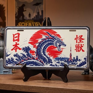 May include: A rectangular metal license plate featuring a graphic illustration of a blue dragon rising from ocean waves against a large red sun. The design includes a city skyline, Mount Fuji, and the Japanese characters 日本 and 怪獣 in red.