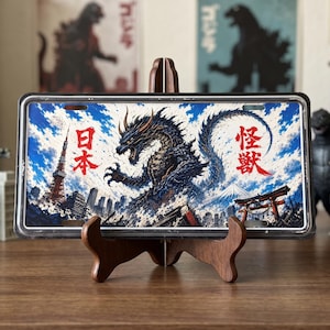 Könnte beinhalten: Ein rechteckiges Metall-Nummernschild mit einer detaillierten Illustration eines dunklen, stacheligen Monsters, das über einer Stadtkulisse mit dem Tokyo Tower und dem Berg Fuji thront. Das Schild zeigt rote japanische Kanji-Zeichen.
