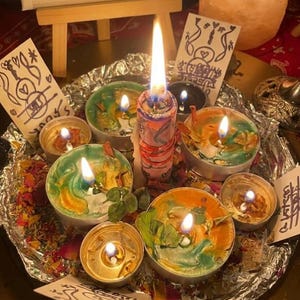 May include: A collection of lit tea light candles and one central pillar candle arranged on a foil-lined tray with scattered dried flower petals. Several small white cards with hand-drawn black ink illustrations are placed among the candles.
