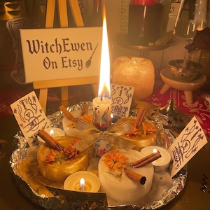 May include: A central lit candle with a gold ribbon, surrounded by smaller white and gold candles, cinnamon sticks, and dried orange flowers on a foil-lined tray. A small wooden easel displays a sign reading WitchEwen On Etsy with a broom icon.