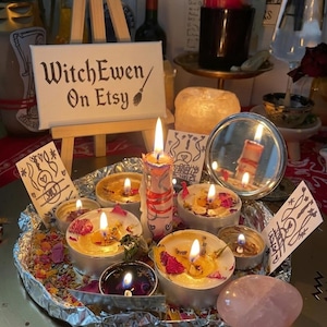 May include: A circular aluminum foil tray holds several lit tea light candles, a taller central candle, scattered dried flower petals, a small round mirror, and a smooth pink stone. A miniature wooden easel displays a sign reading WitchEwen On Etsy.