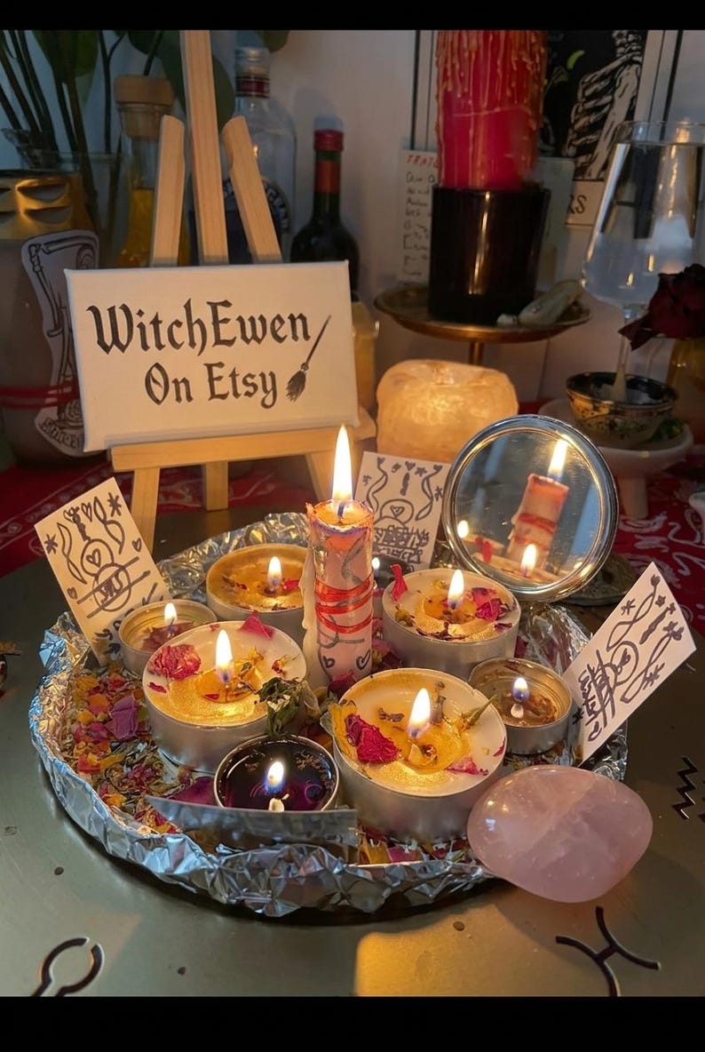 May include: A circular aluminum foil tray holds several lit tea light candles, a taller central candle, scattered dried flower petals, a small round mirror, and a smooth pink stone. A miniature wooden easel displays a sign reading WitchEwen On Etsy.