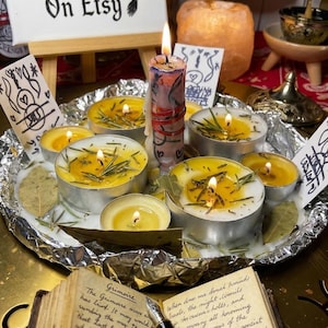 May include: A collection of tea light candles with yellow wax and rosemary sprigs, arranged on a foil-lined tray with a central pillar candle. An open book with handwritten text and a fountain pen sit in the foreground, with a sign reading WitchEwen On Etsy.