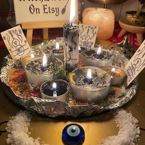 May include: A collection of black tea light candles and one tall pillar candle burning on a foil-lined tray, surrounded by dried flower petals and salt. A blue glass evil eye bead sits in front, with a sign reading WitchEwen On Etsy.