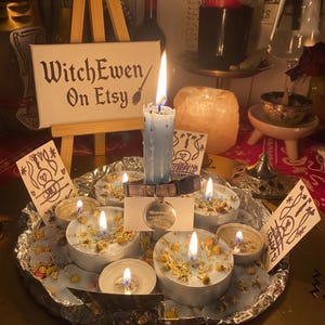 May include: A light blue taper candle and several tea light candles in a foil-lined tray, decorated with dried flowers. A small collar with a round metal tag is wrapped around the taper candle. A wooden easel displays a sign reading WitchEwen On Etsy.