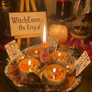 May include: A collection of lit orange tea light candles and a central pillar candle arranged on a foil-lined tray with a small metal bell, dried petals, and hand-drawn cards. A wooden easel displays a sign reading WitchEwen On Etsy.