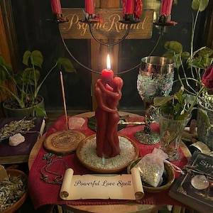 Puede incluir: Una vela roja encendida con la forma de dos figuras abrazadas está sobre una mesa de madera. La mesa está decorada con cristales, un pergamino que dice "Powerful Love Spell" y otros objetos rituales. Un candelabro en forma de cruz está en el fondo.