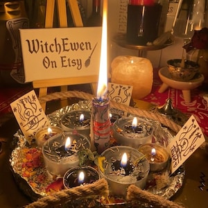 Op de afbeelding: Een rond dienblad bekleed met aluminiumfolie bevat verschillende brandende theelichtjes en een centrale stompkaars met een hoge vlam. Een kleine houten ezel toont een wit bordje met de tekst WitchEwen On Etsy. Gedroogde bloemblaadjes en touwaccenten omringen de kaarsen, met kleine handgetekende kaartjes met lijntekeningen tussen de kaarsen geplaatst.
