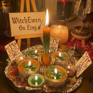 May include: A collection of lit candles in metal tins and a central taper candle on a foil-lined tray, accompanied by a horseshoe, a die, and small cards with hand-drawn symbols. A wooden easel displays a sign reading WitchEwen On Etsy.
