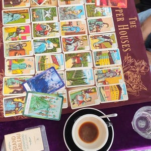 Puede incluir: Un conjunto de cartas de tarot con diversas ilustraciones, dispuestas sobre una superficie burdeos. Las cartas presentan im&aacute;genes de personas, edificios y escenas. Una taza de caf&eacute;, un vaso de agua y un libro tambi&eacute;n est&aacute;n presentes.