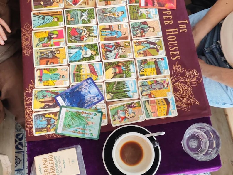 Puede incluir: Un conjunto de cartas de tarot con diversas ilustraciones, dispuestas sobre una superficie burdeos. Las cartas presentan im&aacute;genes de personas, edificios y escenas. Una taza de caf&eacute;, un vaso de agua y un libro tambi&eacute;n est&aacute;n presentes.