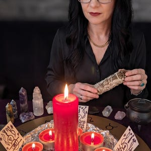 May include: A large red pillar candle surrounded by six smaller red tea light candles on a circular tray covered in aluminum foil. The arrangement includes several small crystals, a small black cauldron, and two white cards with black ink drawings.