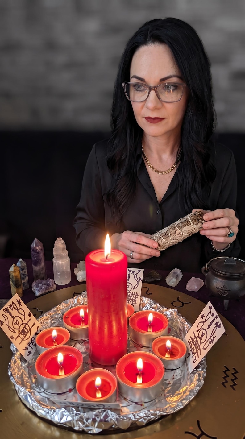 May include: A large red pillar candle surrounded by six smaller red tea light candles on a circular tray covered in aluminum foil. The arrangement includes several small crystals, a small black cauldron, and two white cards with black ink drawings.