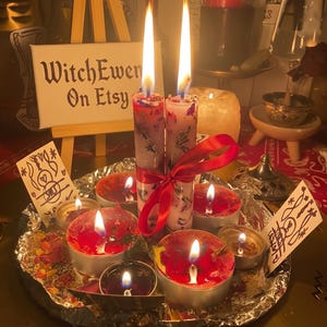 Puede incluir: Dos velas blancas altas con pétalos de flores secas incrustados, atadas con una cinta roja, se encuentran en el centro de una bandeja forrada con papel de aluminio, rodeadas de varias velas de té rojas encendidas y dos tarjetas con dibujos.