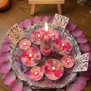Könnte beinhalten: Ein silbernes Alufolientablett gefüllt mit rosa Rosenblättern, Lavendelzweigen und mehreren brennenden Teelichtern, die eine zentrale Stumpenkerze umgeben. Drei kleine weiße Karten mit handgezeichneten schwarzen Tintensymbolen liegen dazwischen.