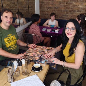 May include: A square board game with a geometric pattern of triangular cards sits on a wooden table between two people. The game features a dark border with gold accents. A menu, a cup of tea, and metal condiment holders are also on the table.