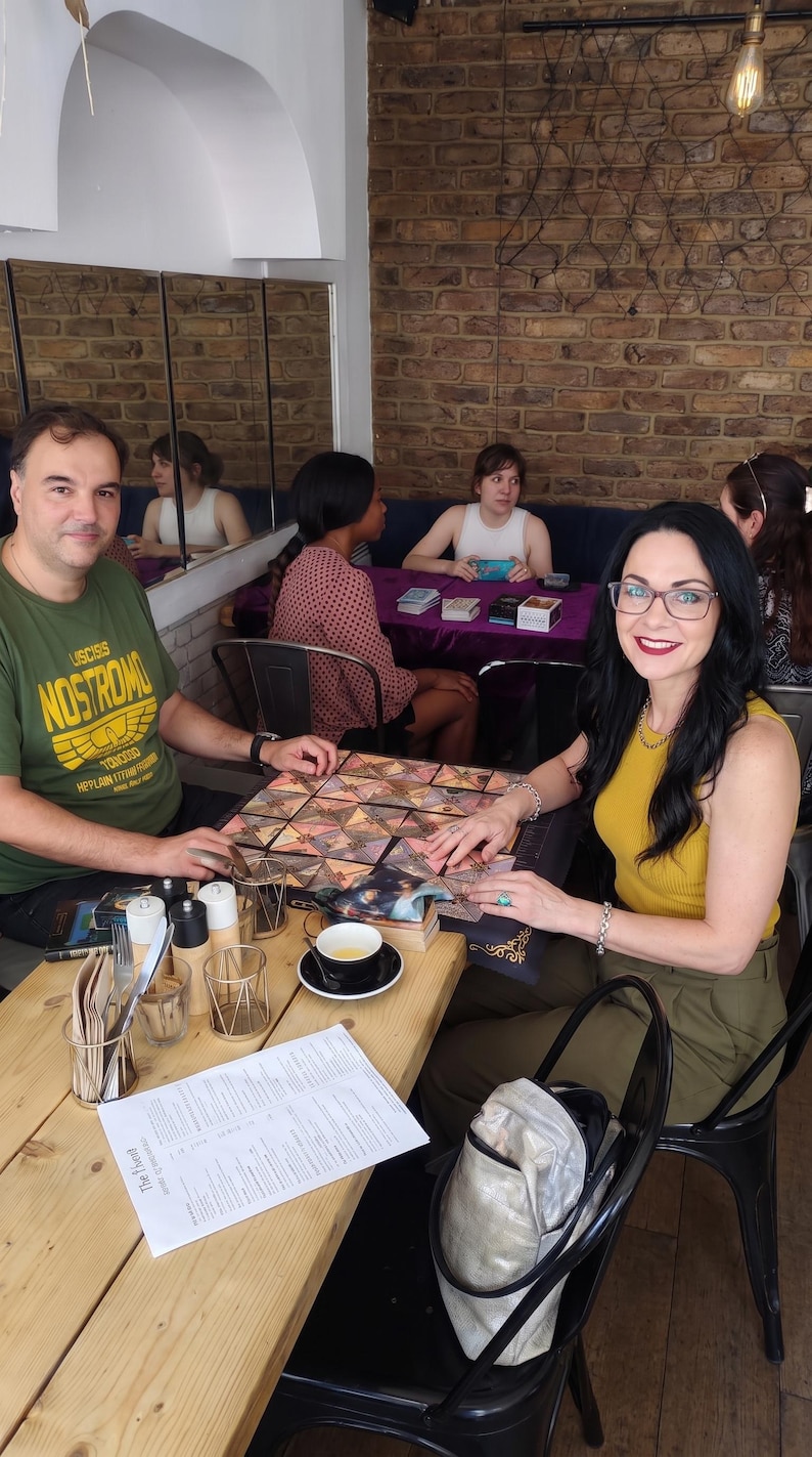 May include: A square board game with a geometric pattern of triangular cards sits on a wooden table between two people. The game features a dark border with gold accents. A menu, a cup of tea, and metal condiment holders are also on the table.