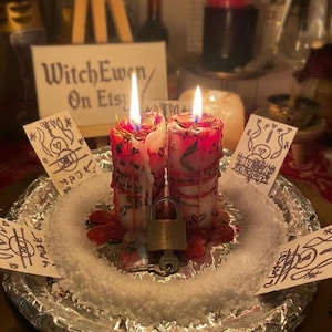 May include: Two white pillar candles with red wax drips, embedded dried herbs, and lit wicks, positioned on a foil-covered plate with white salt. A metal padlock with a key sits between the candles, surrounded by four small cards with hand-drawn symbols.
