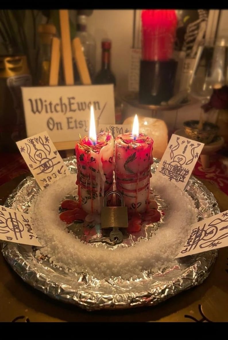 May include: Two white pillar candles with red wax drips, embedded dried herbs, and lit wicks, positioned on a foil-covered plate with white salt. A metal padlock with a key sits between the candles, surrounded by four small cards with hand-drawn symbols.