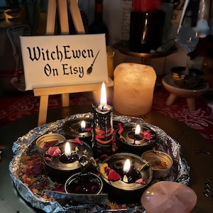 May include: A small wooden easel displays a white sign with the black text WitchEwen On Etsy and a broom icon. Below, a circular tray lined with aluminum foil holds several black tea light candles, dried flower petals, and a pink crystal.